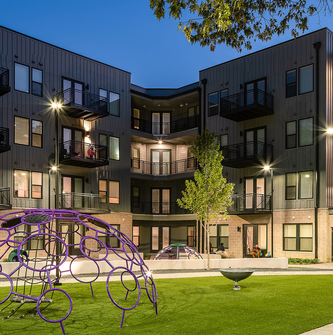 Evening courtyard of an apartment complex featuring a modern playground structure, neatly manicured turf, and soft outdoor lighting. Residents sit and relax near the building entrance.
