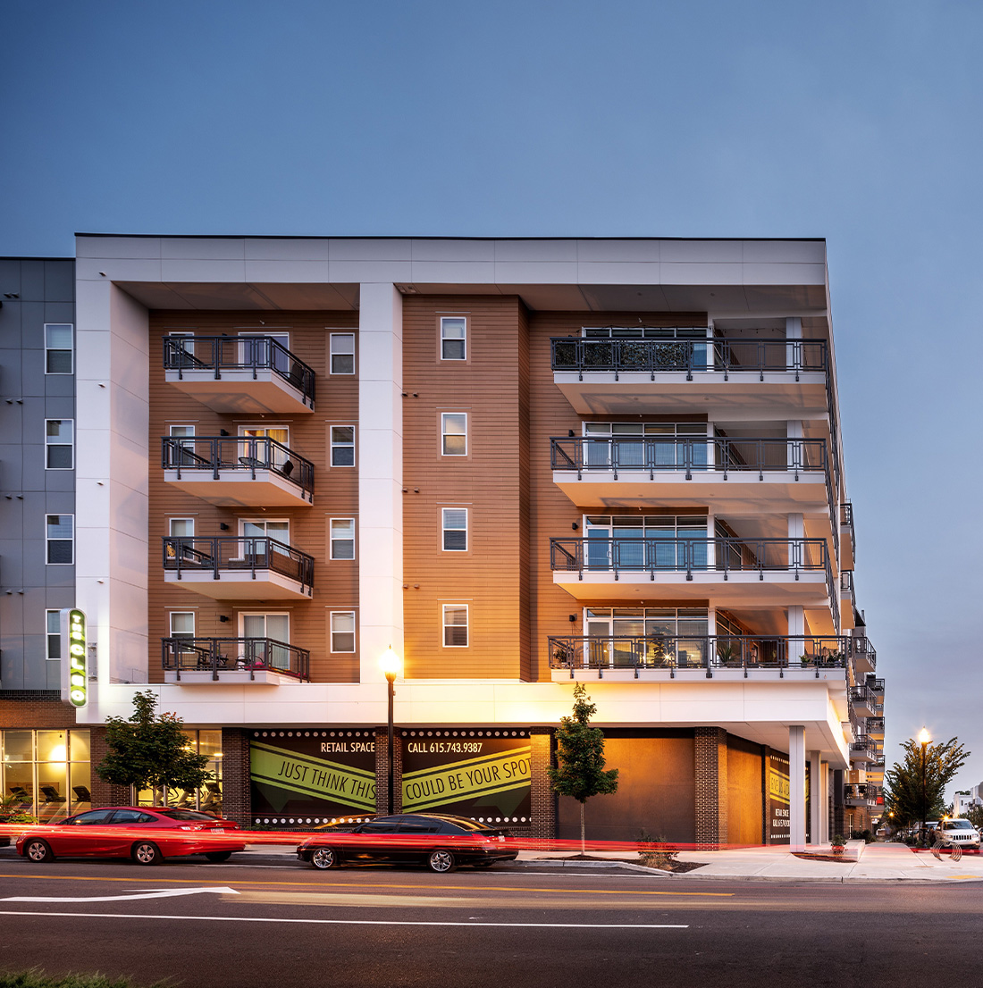 Street-level view of a corner apartment building with prominent balconies, wood-paneled siding, and retail space banners reading “Just think... this could be your spot.