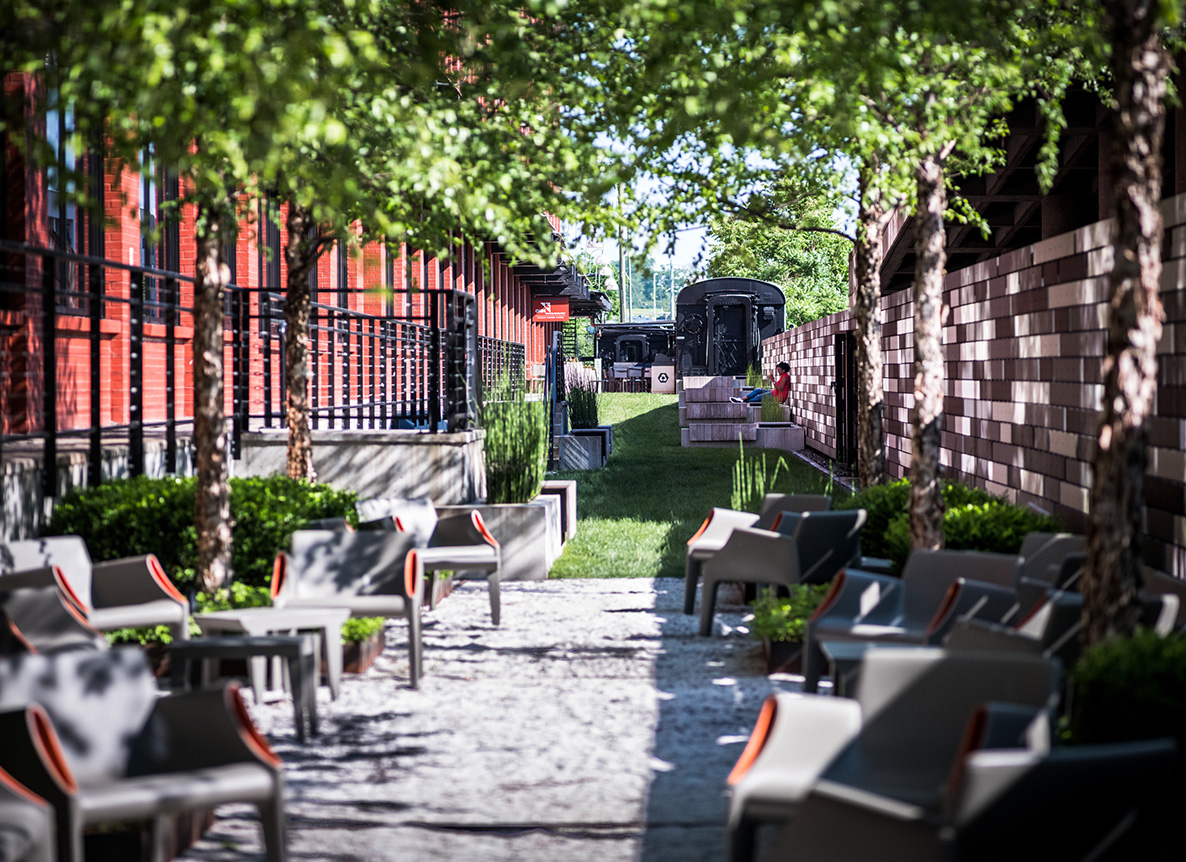 A modern outdoor seating area is nestled between a red brick building and a patterned stone wall, lined with trees and greenery. Contemporary gray chairs with orange accents are arranged along a gravel path that leads to a grassy section with vintage black train cars in the distance. A person in red sits near the train cars, adding a pop of color to this urban, park-like setting.