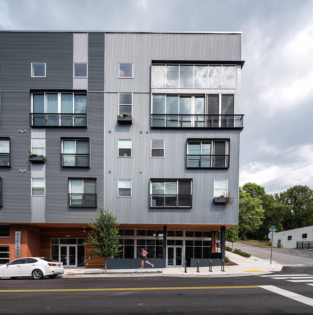 A modern mixed-use building with large windows, textured metal façade, and street-level retail sits at the corner of an urban intersection.