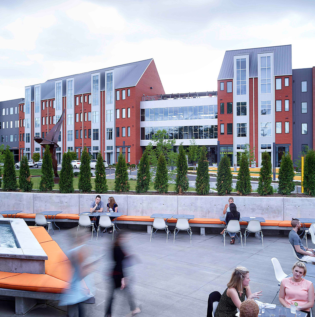Outdoor patio with orange bench seating, modern tables and chairs, and people socializing in front of a backdrop of red brick apartment buildings and sculptural landscaping.