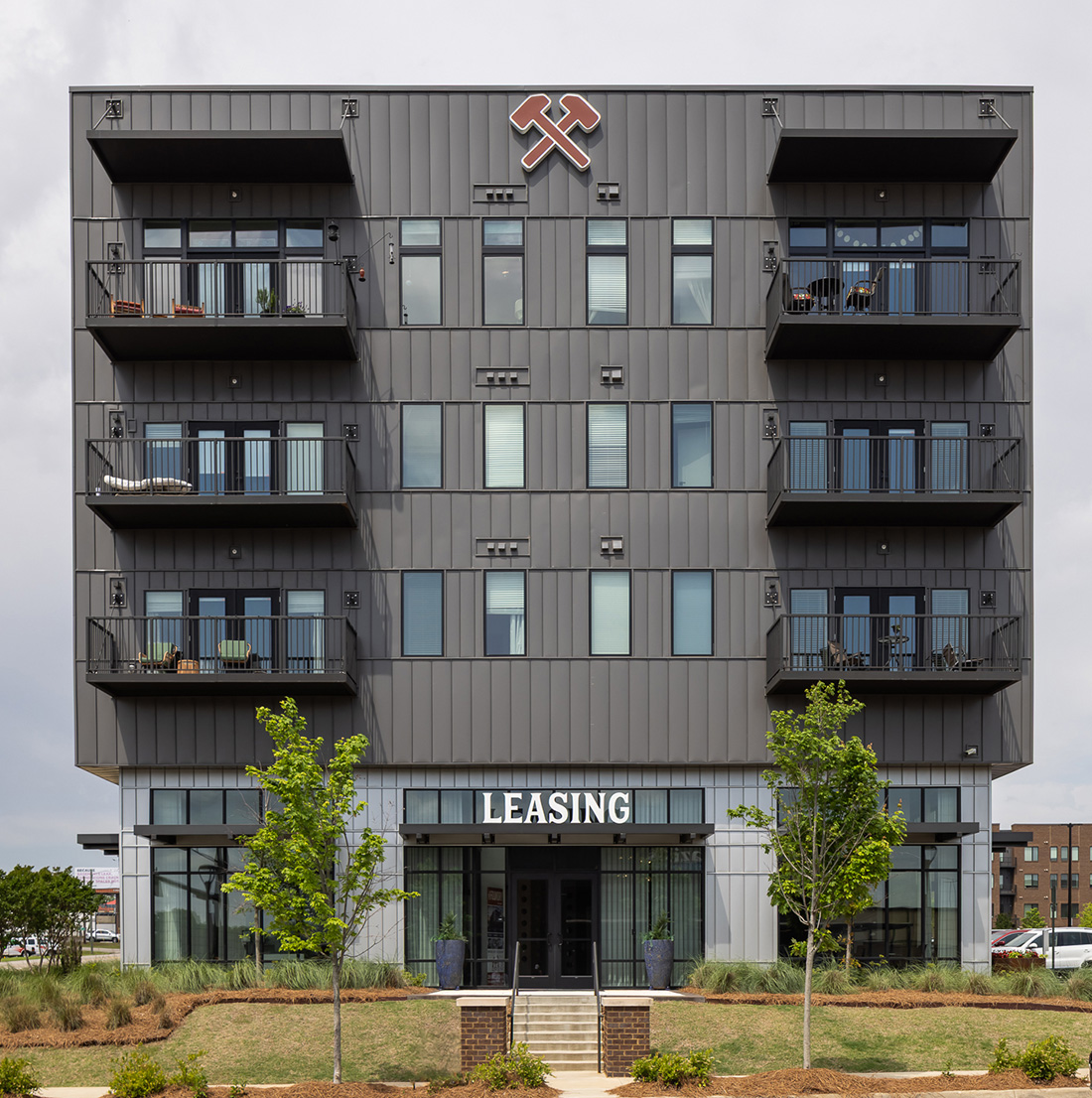 Front view of a modern mid-rise apartment building with dark metal siding and private balconies. The entrance features large glass doors under a prominent “LEASING” sign, flanked by small landscaped trees and minimal brick steps. A crossed-hammer logo is displayed above the top-floor windows.