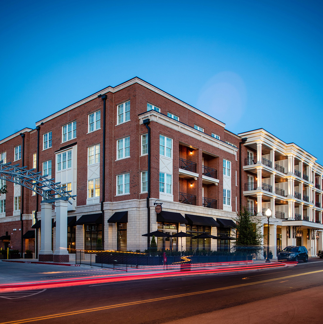 Evening street view of a vibrant mixed-use development with ground-level retail, upper-story apartments, brick and stone façade, glowing windows, and light trails from passing cars under a deep blue sky.