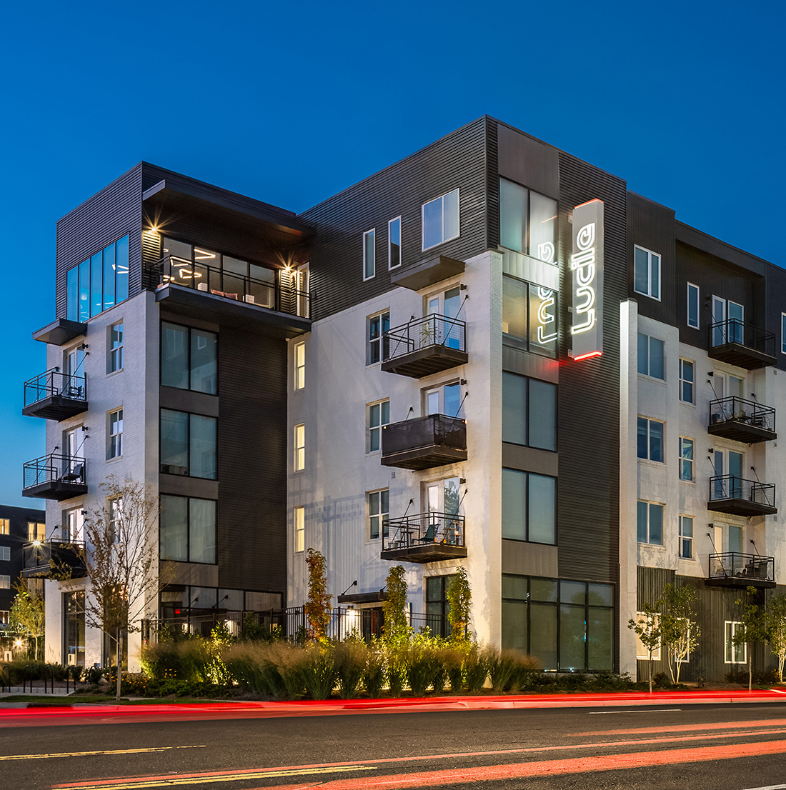 A five-story residential building features modern balconies, large windows, and glowing signage, with landscaped surroundings and a lit street at night.