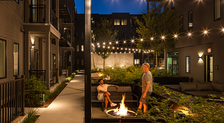 Cozy nighttime courtyard scene with string lights, lush greenery, and two residents relaxing by a fire pit in a modern apartment complex.