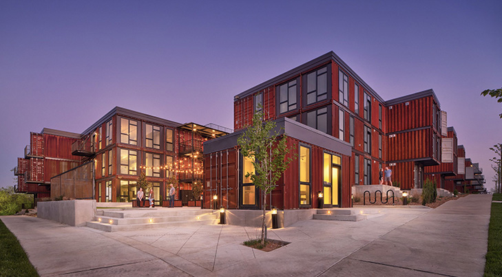 Bold modern apartment complex made from stacked red shipping containers, glowing warmly at dusk with lit walkways, glass walls, and industrial charm.