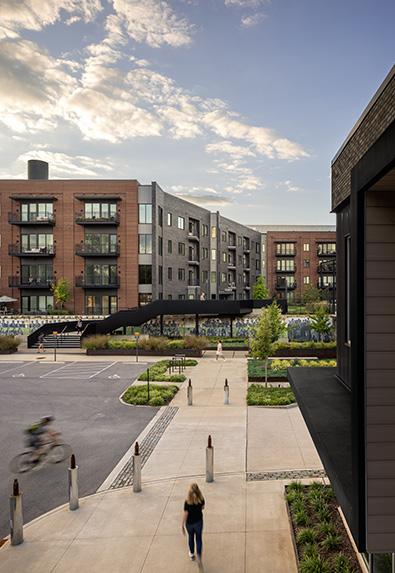 Elevated view of a pedestrian-friendly apartment complex featuring red and gray brick buildings, landscaped paths, and a black metal bridge connecting two areas.