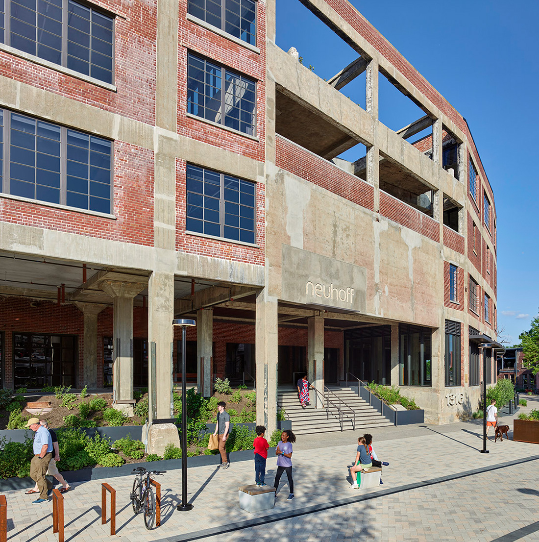 Street-level view of the "Neuhoff" building entrance, showcasing partially preserved brick-and-concrete façade, wide steps, and active public space. People and children interact in the modern plaza surrounded by greenery and restored industrial architecture.