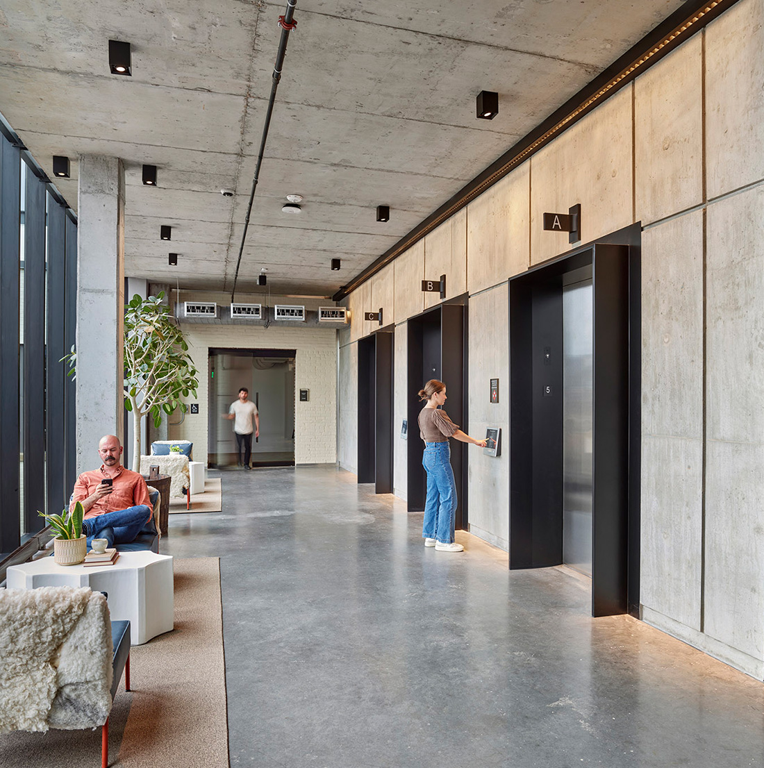 A modern elevator lobby with an industrial design features concrete walls and ceilings, exposed pipes, and sleek black elevator doors labeled A and B. A woman is pressing the elevator button while another person walks down the hallway. On the left, a man sits in a cozy seating area with sheepskin-covered chairs, white side tables, and potted plants near large floor-to-ceiling windows that let in natural light.