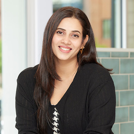Woman with long dark hair wearing a black cardigan over a patterned top, smiling and seated with arms crossed on a counter, indoors with large windows behind.