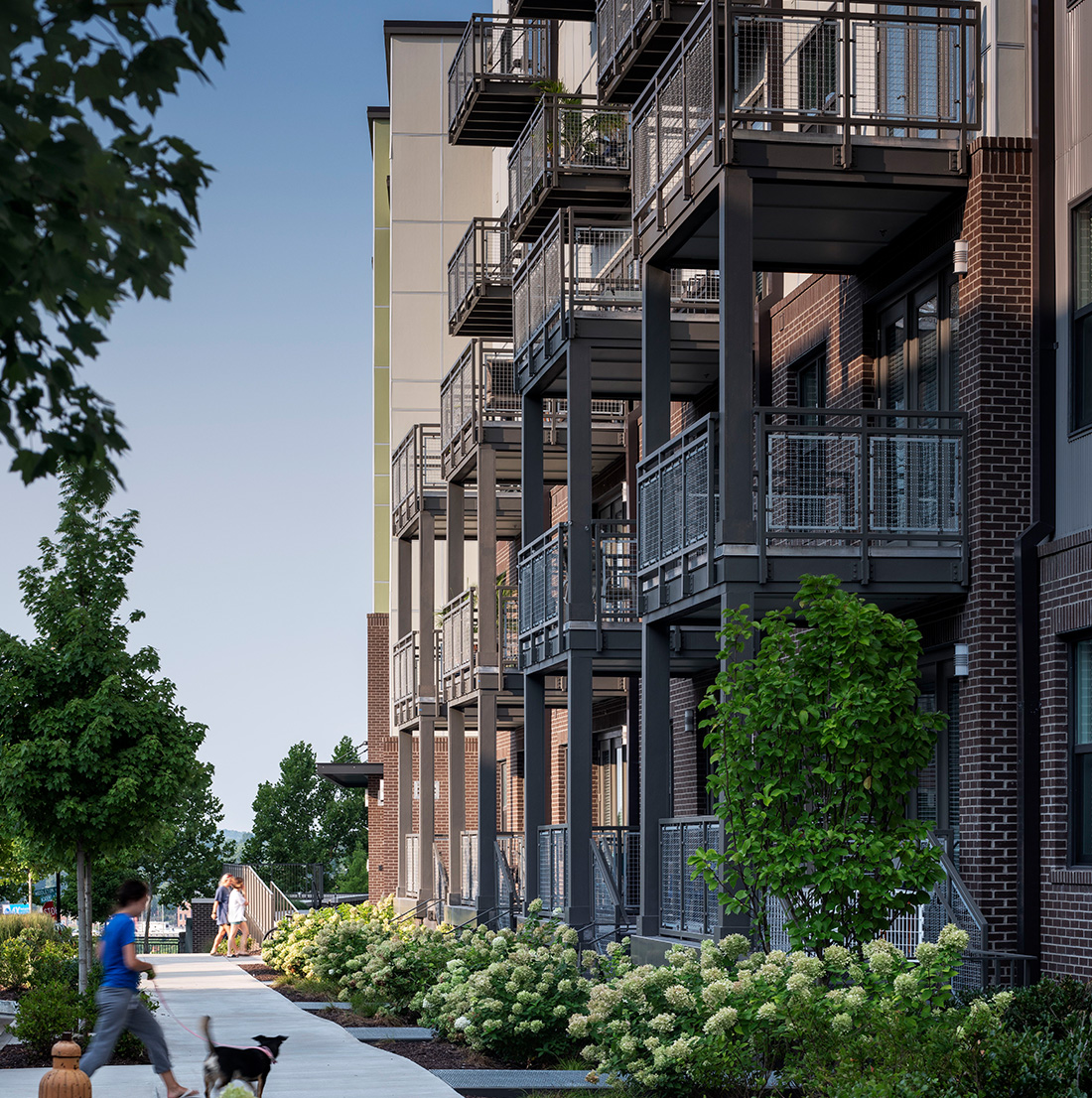 Sidewalk along the apartment buildings showing several private balconies, manicured landscaping, and residents walking dogs and chatting.