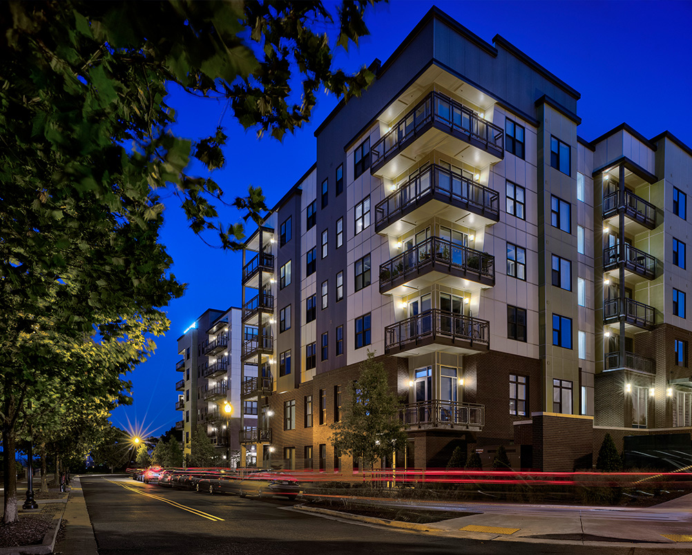 Nighttime street-level view of a modern apartment building with balconies and lit windows, framed by trees and light trails from passing cars.