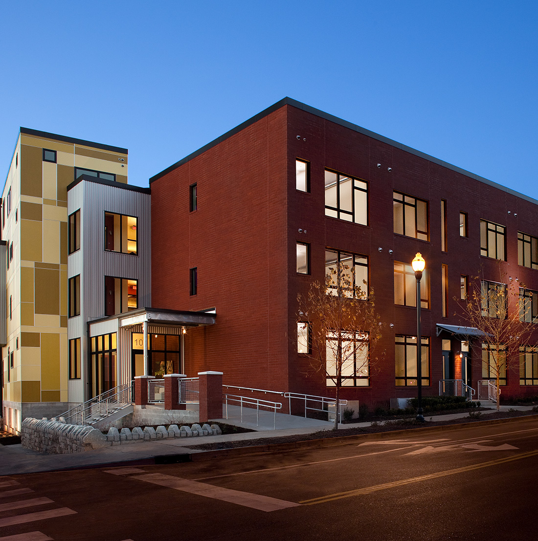Corner view of a contemporary apartment complex with red brick and yellow paneling, featuring accessible ramps and well-lit pathways during evening hours.