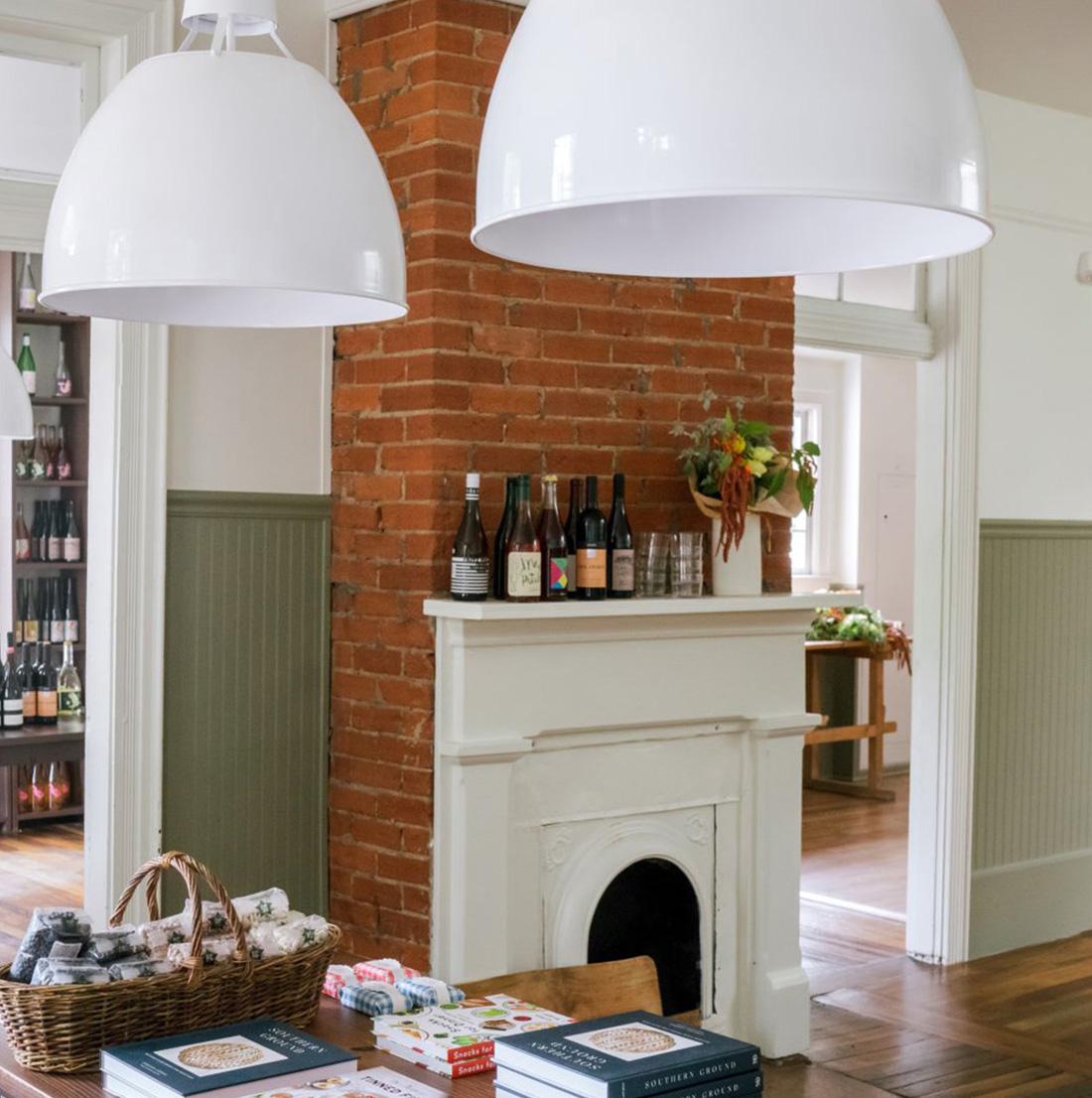 Interior shot with a red brick fireplace, white mantle, wine bottles, and a table displaying books, soaps, and gourmet goods under large white pendant lights.