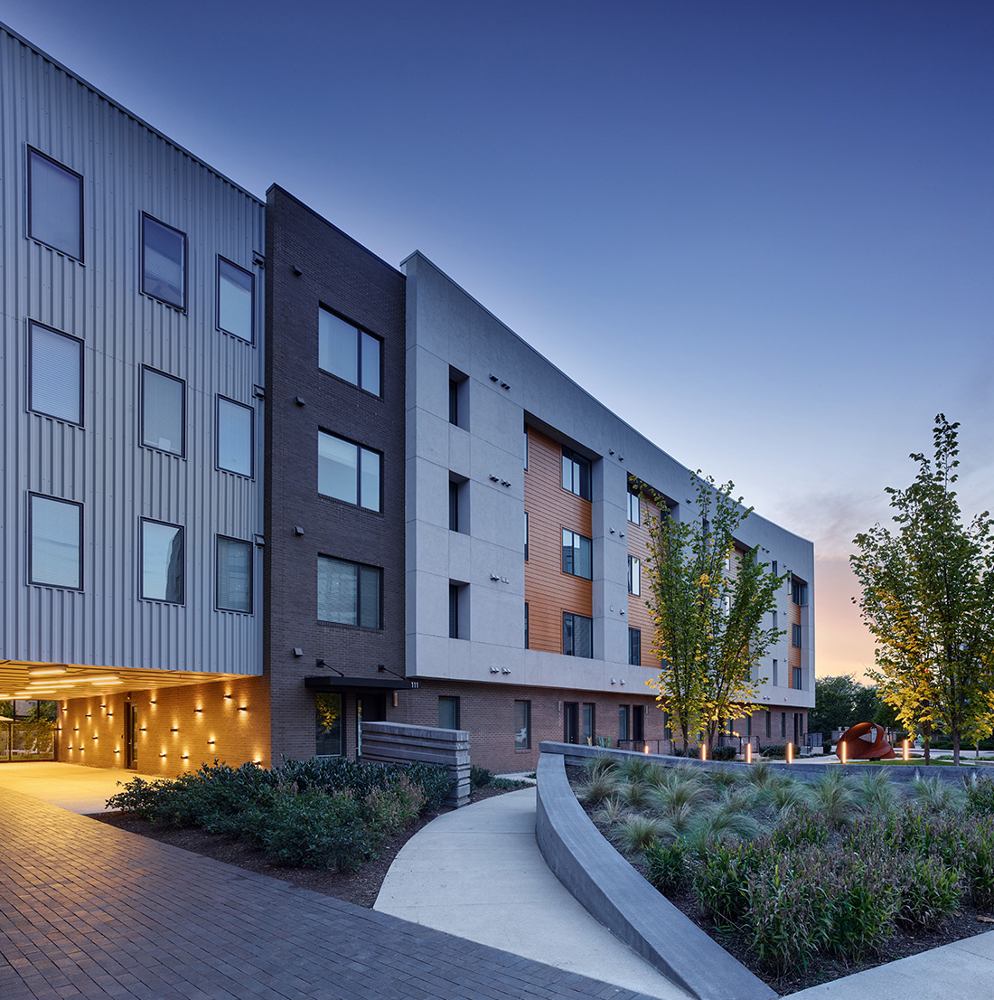 A multi-story apartment building with mixed exterior finishes is shown at dusk with landscaped pathways and outdoor lighting.