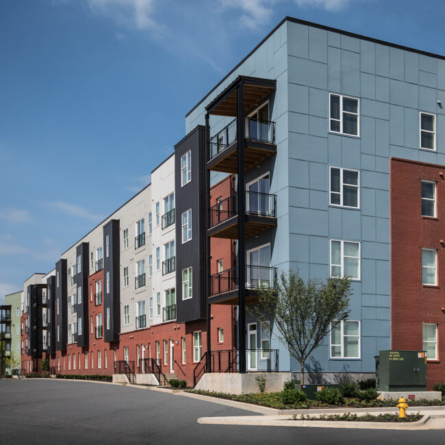 Modern four-story apartment building with a mix of red brick and blue-gray panel siding, accented by balconies with black metal railings and a clean urban streetscape.