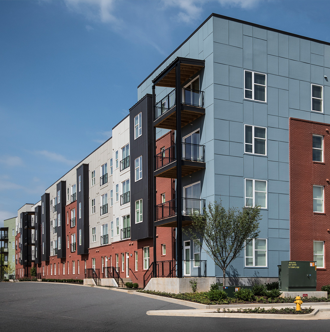 Modern four-story apartment building with a mix of red brick and blue-gray panel siding, accented by balconies with black metal railings and a clean urban streetscape.