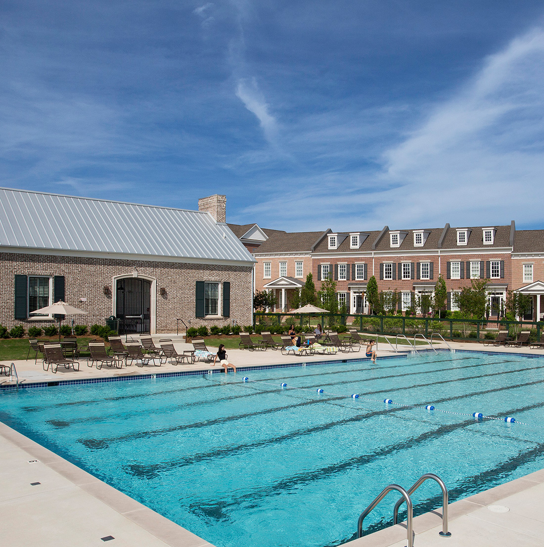 Large rectangular swimming pool with designated swim lanes, surrounded by lounge chairs and umbrellas. In the background, a brick clubhouse with a metal roof and adjacent townhouses with dormer windows provide a resort-style setting.