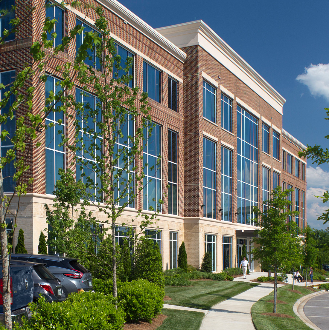 Angled view of a red brick office building featuring large glass windows. The building is bordered by newly planted trees and a landscaped sidewalk.