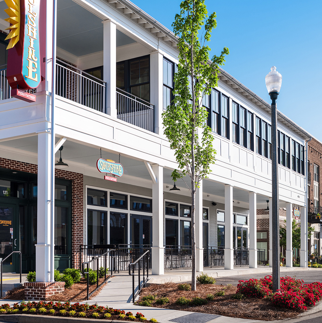 Modern two-story retail building with white trim and large storefront windows, featuring the Soulshine Pizza Factory. The building includes shaded walkways and is surrounded by landscaped flowers and mature trees.