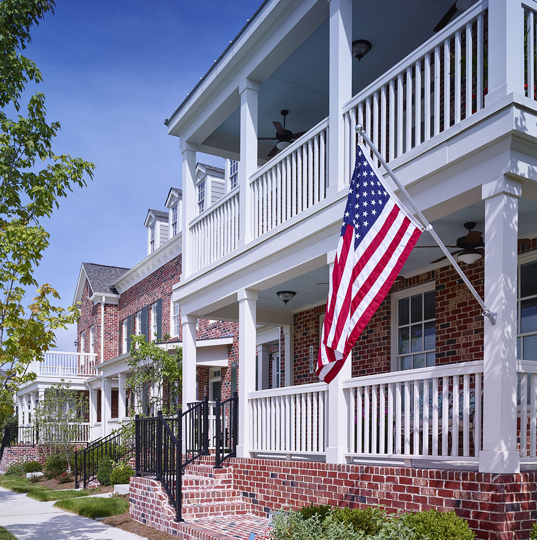 Close-up view of a row of brick townhomes featuring double front porches with white railings. An American flag is displayed on one of the porches, and ceiling fans are visible overhead. Trees line the walkway in front of the homes.