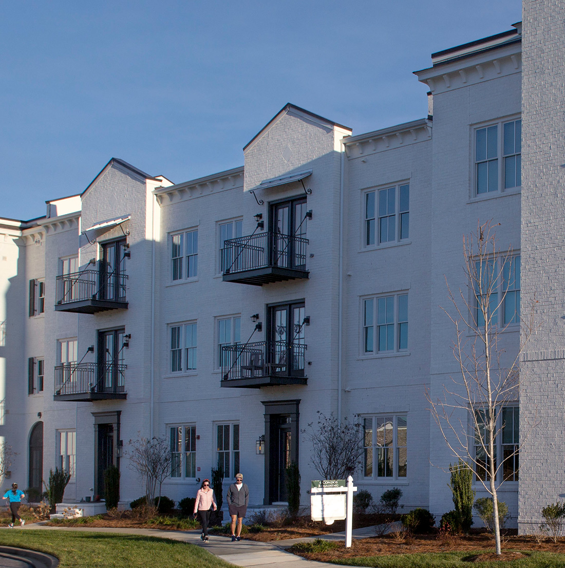 Side view of a white brick residential building featuring multiple black iron balconies and tall windows. Several people are walking along the sidewalk in front of the landscaped area bordering the building.