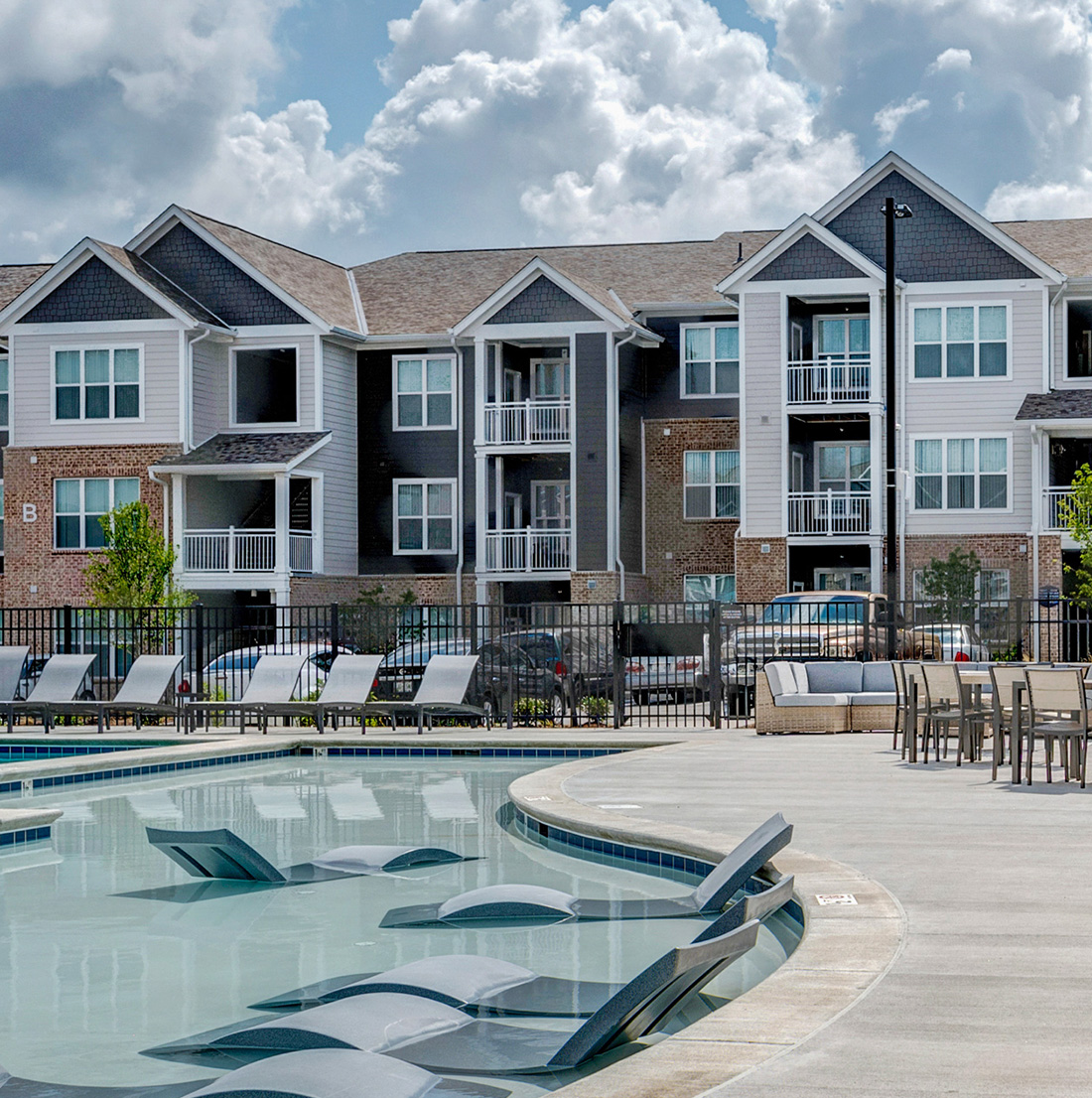 Curved resort-style swimming pool with built-in lounge chairs and surrounding deck seating. The fenced pool area is situated in front of three-story apartment buildings featuring brick and siding exteriors, beneath a partly cloudy blue sky.