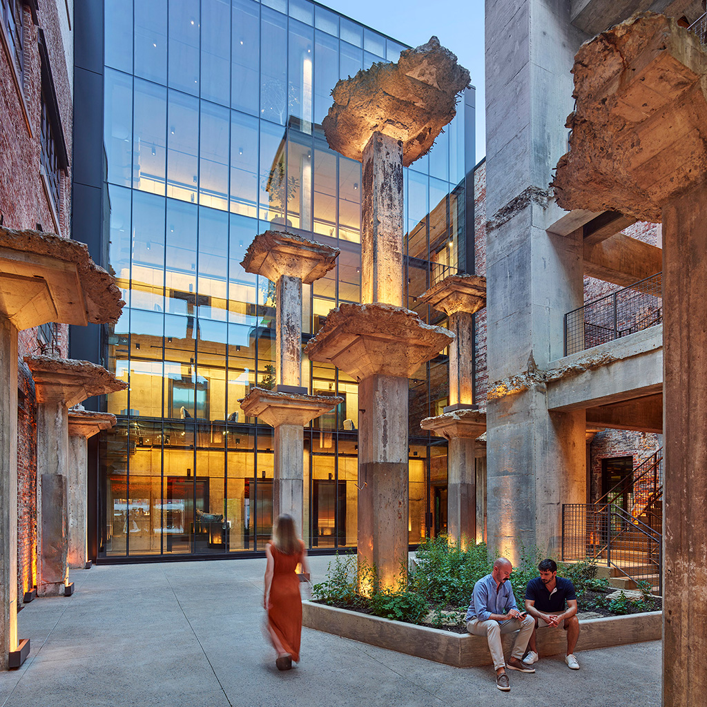 Outdoor courtyard with exposed concrete columns and modern glass building in the background, two men seated on a planter bench having a conversation while a woman in a dress walks by.