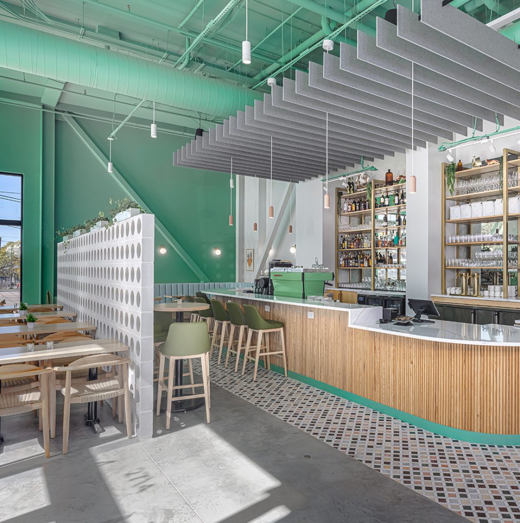 A modern cafe interior with mint green walls, wood and marble finishes, suspended acoustic panels, and a well-stocked bar.