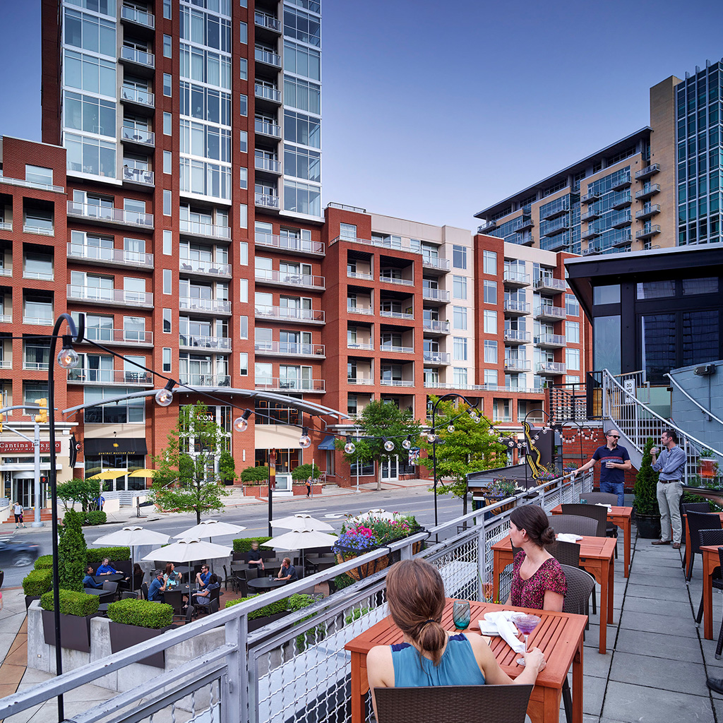 Urban outdoor dining terrace overlooking a city street with apartment buildings, patrons seated at tables, and people conversing on a staircase.