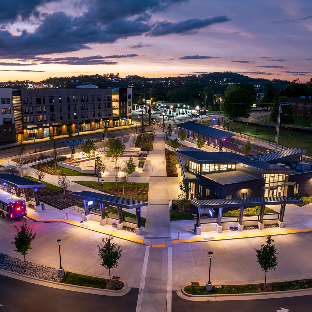 An illuminated transit center at dusk featuring modern bus shelters, landscaped walkways, and surrounding urban development under a dramatic evening sky.
