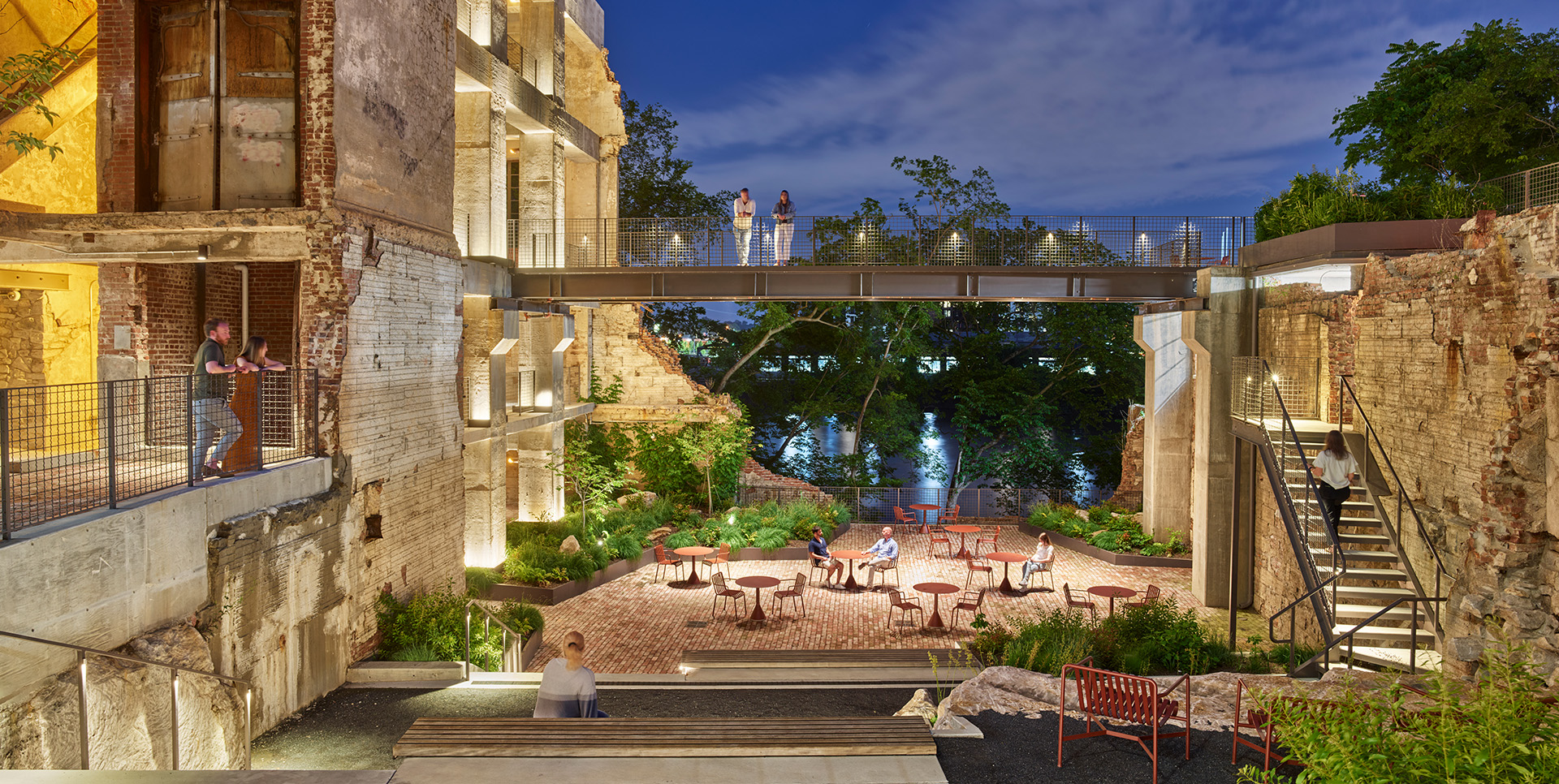 Evening view of an outdoor courtyard surrounded by historic brick ruins with modern landscaping and seating, including several people seated and standing, under ambient lighting.