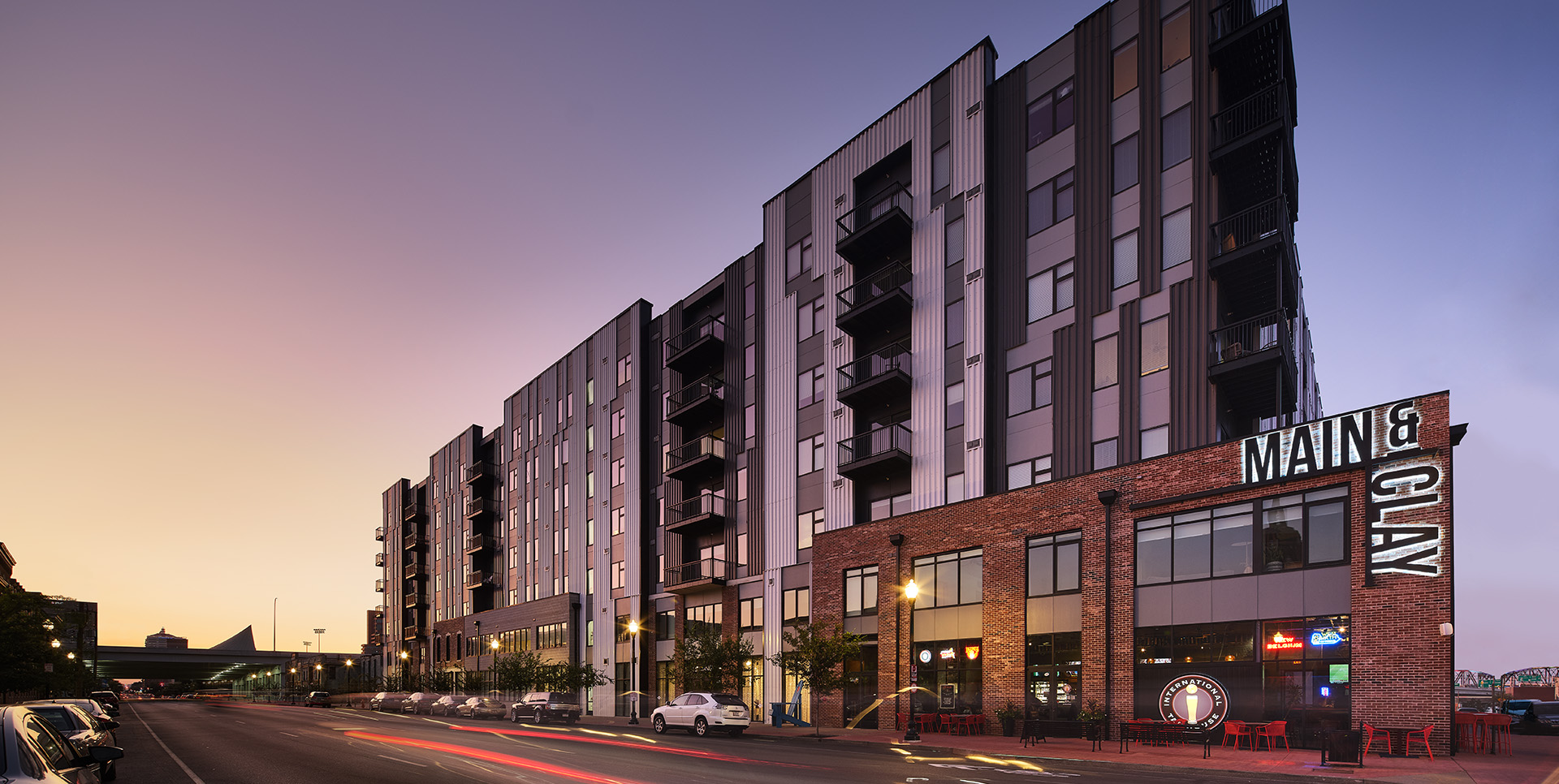Wide-angle exterior shot of a tall, modern apartment building named "Main & Clay," with street-level retail spaces, parked cars, and a softly lit urban environment at dusk.