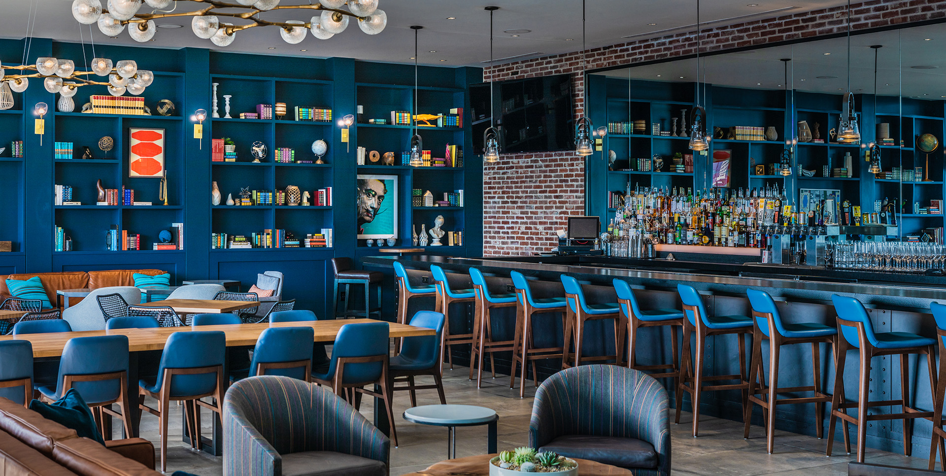 Modern lounge and bar area with deep blue bookshelves, wooden tables, cushioned seating, and ambient lighting fixtures.