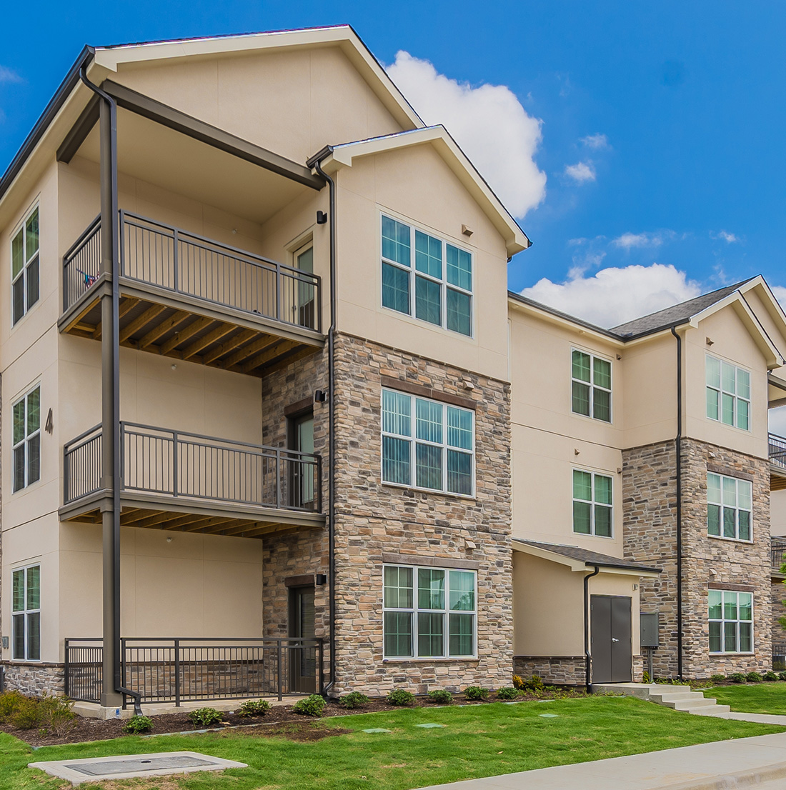A three-story residential building with beige siding, stone accents, and private balconies is shown on a sunny day with neatly maintained landscaping.