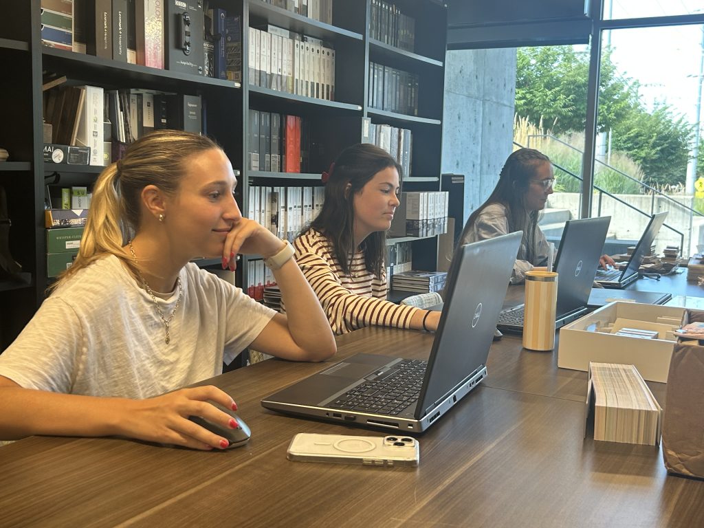 Three women sit side by side at a long wooden table, each working on a laptop in a modern office space. Shelves filled with books and samples line the back wall, while large windows let in natural light and offer a view of greenery outside. A smartphone, mouse, coffee tumbler, and sample boxes are on the table, contributing to the workspace's organized yet casual vibe.