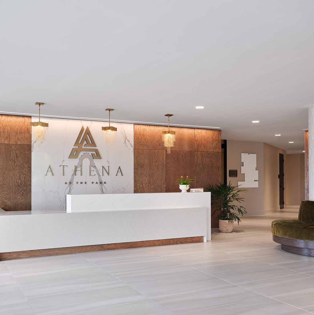 A sleek and modern reception area features a minimalist white front desk set against a backdrop of warm wood paneling and white marble with gold accents. The wall behind the desk displays a large metallic logo reading "ATHENA AT THE PARK." Elegant pendant lights hang from the ceiling, and potted plants add a touch of greenery to the clean, sophisticated space.