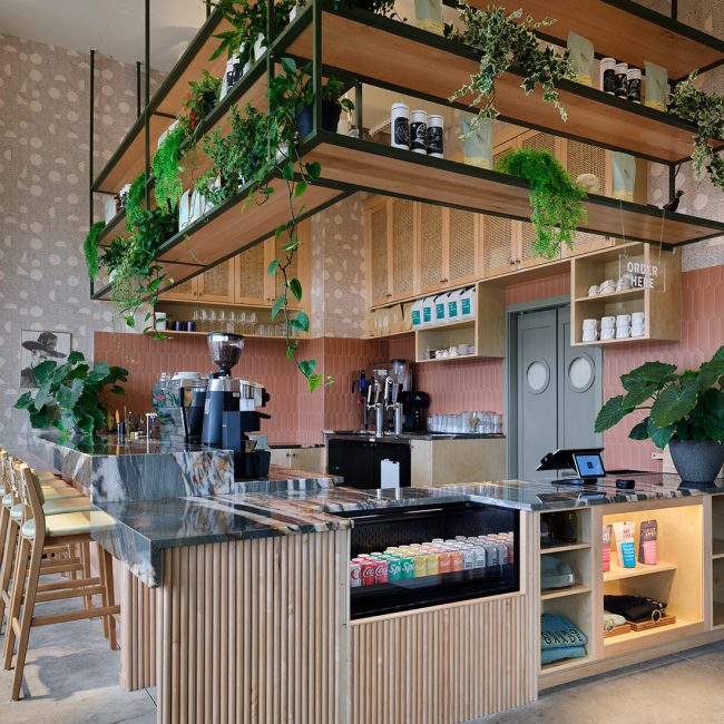 A stylish, modern coffee bar features a curved marble countertop, wood accents, and suspended shelving filled with plants and coffee products. The space has a warm, inviting atmosphere with soft pink tile walls, built-in shelving, and natural lighting.