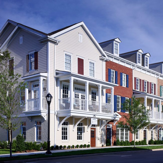 Row of elegant, multi-story townhomes with a mix of brick and siding exteriors in neutral tones. Each unit features white-trimmed windows, decorative shutters, and upper-level balconies with white railings. The buildings have pitched roofs with dormer windows and are framed by neatly landscaped greenery and a tree-lined sidewalk under a bright blue sky.