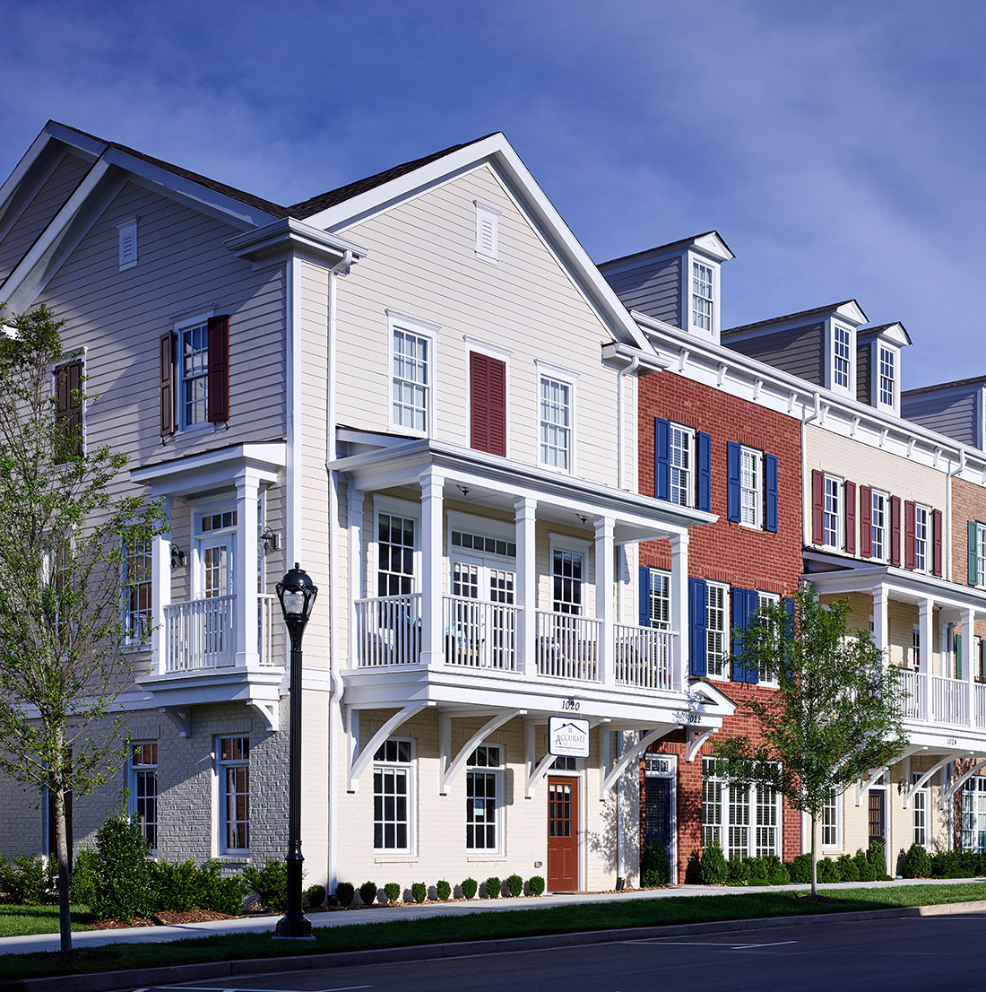 Row of elegant, multi-story townhomes with a mix of brick and siding exteriors in neutral tones. Each unit features white-trimmed windows, decorative shutters, and upper-level balconies with white railings. The buildings have pitched roofs with dormer windows and are framed by neatly landscaped greenery and a tree-lined sidewalk under a bright blue sky.