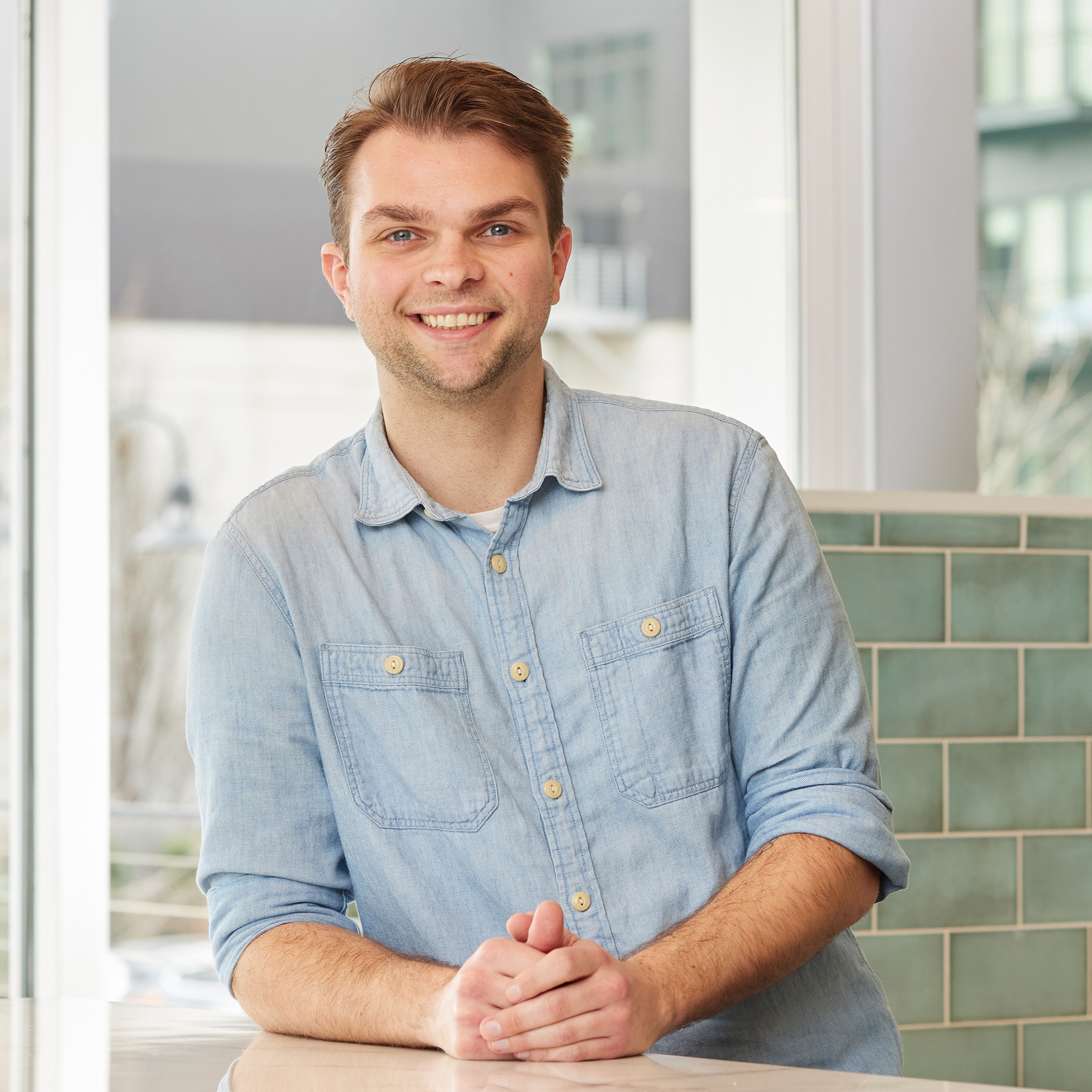 Smiling man in a light blue button-down shirt leaning forward on a counter in a modern office environment.