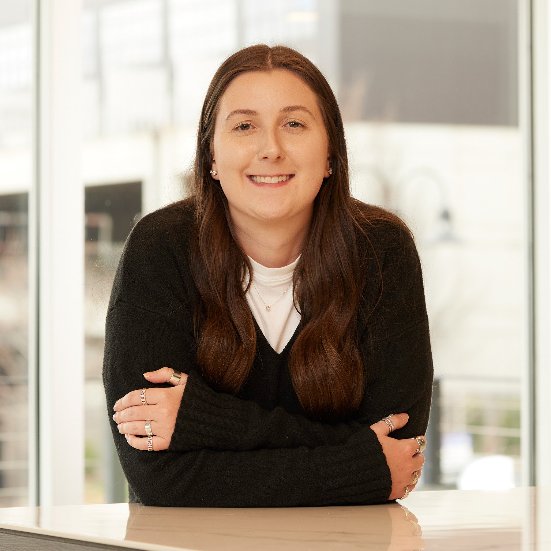 Smiling woman with long brown hair in a black sweater leaning on a counter in a bright office setting.