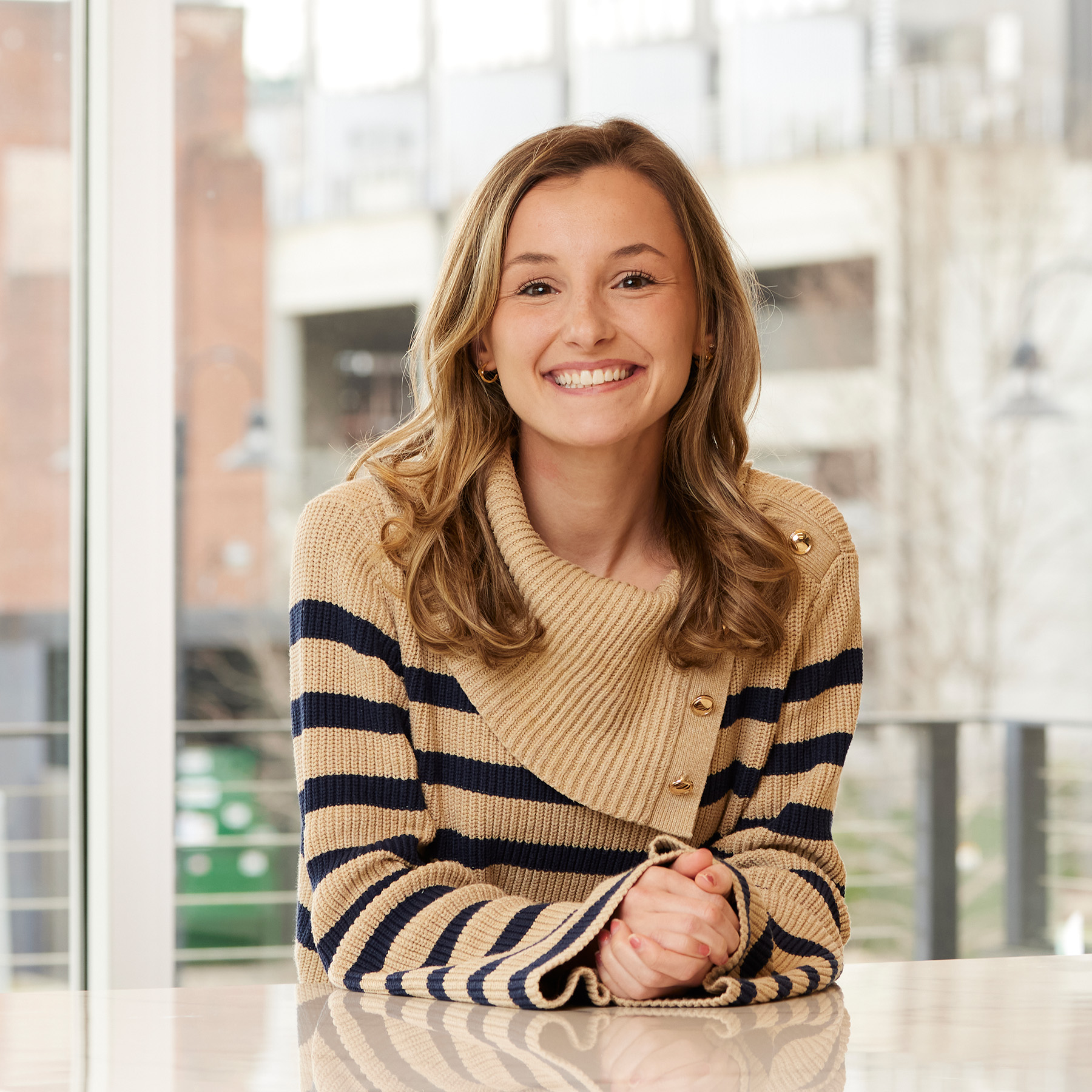 Smiling woman in a tan and navy striped sweater sitting at a counter in a light-filled interior space.