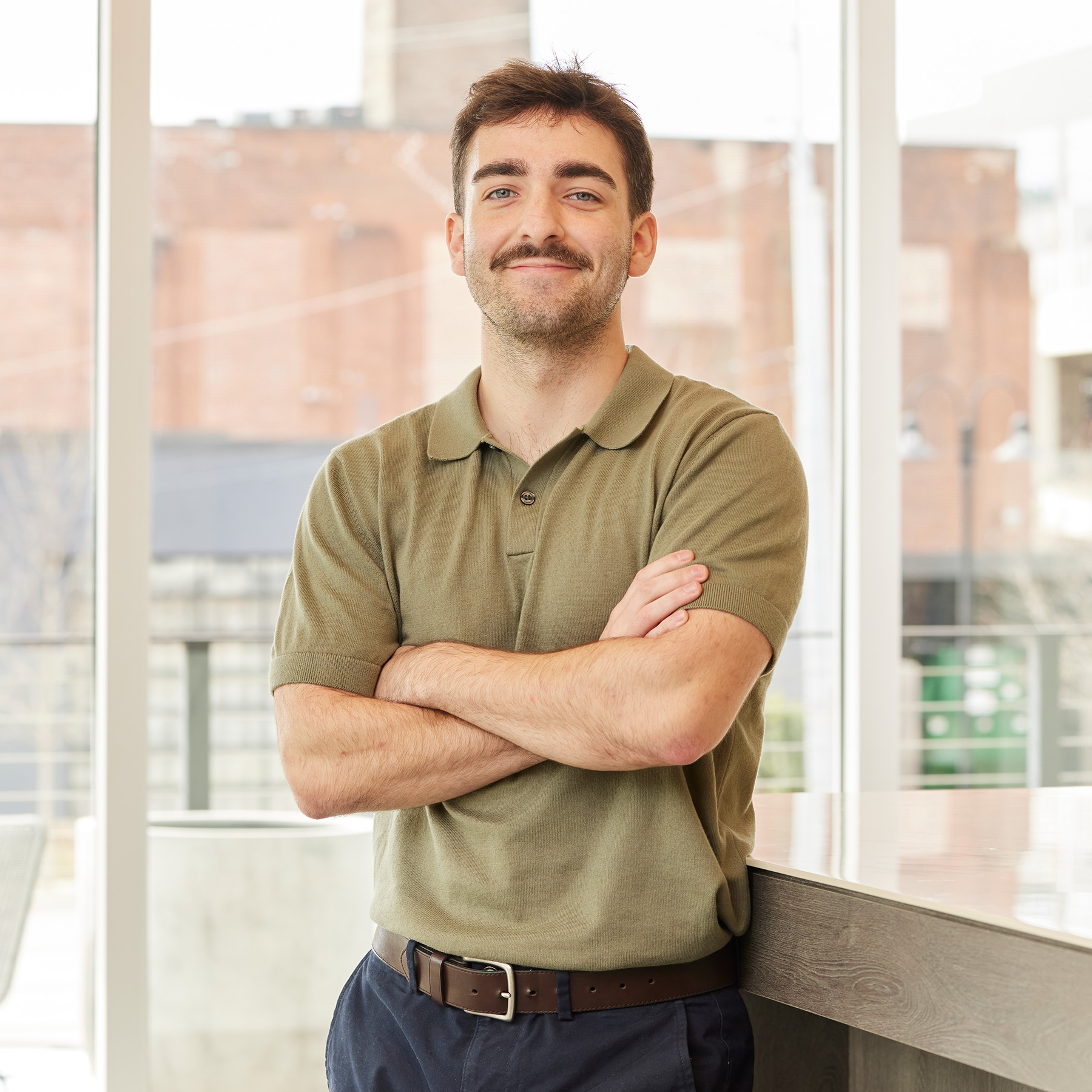 Man with a mustache wearing a green polo shirt standing with arms crossed in a bright indoor space.